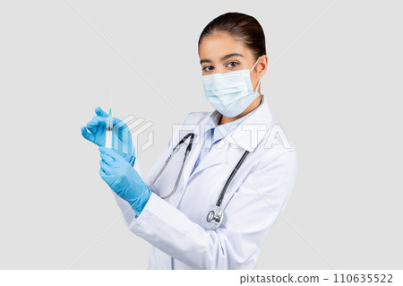 A focused female doctor in a lab coat and surgical mask holds a syringe A focused female doctor in a lab coat and surgical mask holds a syringe 110635522
