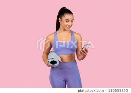 Smiling caucasian lady in sportswear holding rolled-up yoga mat and checking her smartphone, ready for workout 110635530