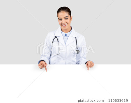 Radiant european doctor with a stethoscope, smiling and pointing downwards towards an empty banner Radiant european doctor with a stethoscope, smiling and pointing downwards towards an empty banner 110635619