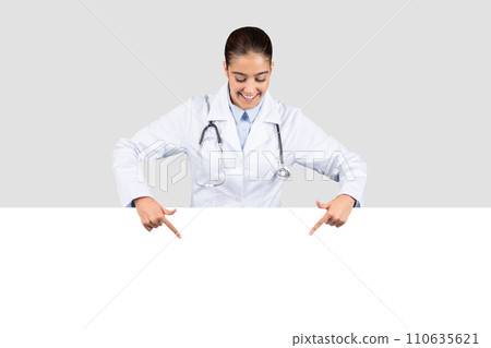 A smiling female doctor in a white coat with a stethoscope points downwards at an empty banner A smiling female doctor in a white coat with a stethoscope points downwards at an empty banner 110635621