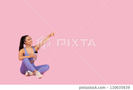 Fitness advertisement. Sporty lady sitting with bottle of water and pointing at copy space over pink background Fitness advertisement. Sporty lady sitting with bottle of water and pointing at copy space over pink background 110635639