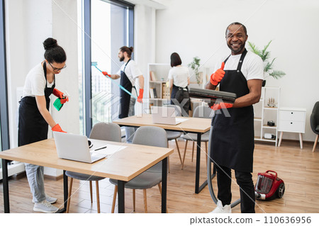 African American man wearing black apron vacuuming floor of modern office. 110636956