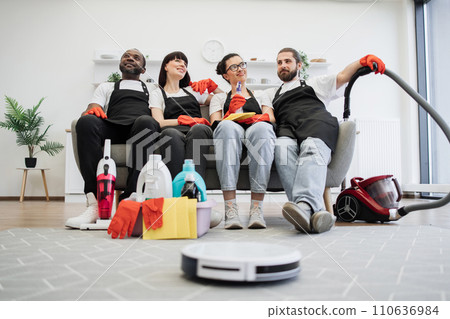 Team of housekeepers sitting on couch while robot vacuum cleaner cleans floor. Team of housekeepers sitting on couch while robot vacuum cleaner cleans floor. 110636984