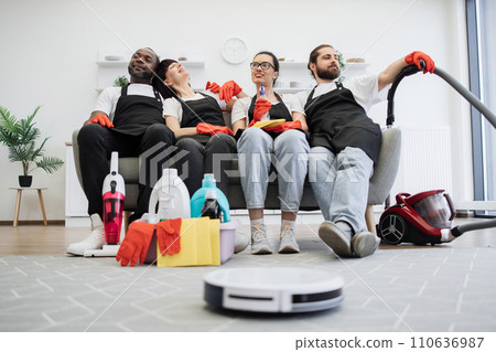 Team of housekeepers sitting on couch while robot vacuum cleaner cleans floor. Team of housekeepers sitting on couch while robot vacuum cleaner cleans floor. 110636987