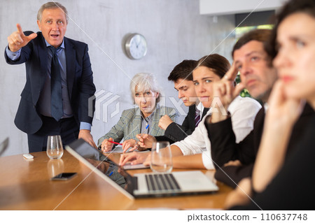 Outraged old manager getting mad at young adult mature office workers sitting with reports and laptops at table in meeting room Outraged old manager getting mad at young adult mature office workers sitting with reports and laptops at table in meeting room 110637748