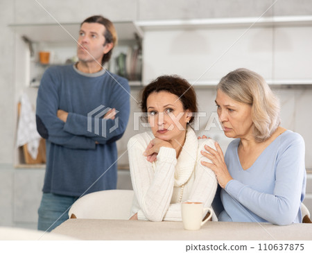 Man is actively quarreling with his thinking wife in kitchen, standing behind her, and elderly mother hugs and calms her daughter Man is actively quarreling with his thinking wife in kitchen, standing behind her, and elderly mother hugs and calms her daughter 110637875