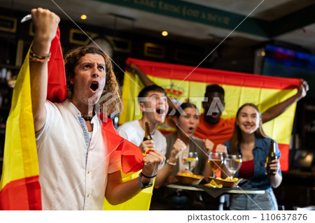 Cheerful multiracial soccer fans waving the flag of Spain while drinking beer in sport bar Cheerful multiracial soccer fans waving the flag of Spain while drinking beer in sport bar 110637876