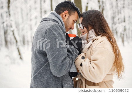 Indian man and caucasian woman walking in winter forest together. Lovely couple hugging and holding a thermos with hot tea. Man wearing grey coat and woman beige coat. Indian man and caucasian woman walking in winter forest together. Lovely couple hugging and holding a thermos with hot tea. Man wearing grey coat and woman beige coat. 110639022