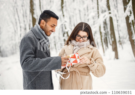 Indian man and caucasian woman walking in winter forest together. Man and woman opening a gift box. Man wearing grey coat and woman beige coat. Indian man and caucasian woman walking in winter forest together. Man and woman opening a gift box. Man wearing grey coat and woman beige coat. 110639031