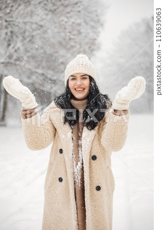 Brunette woman standing at forest and posing for a photo. Woman glad to see snow. Girl wearing winter clothes and knitted hat with gloves. Brunette woman standing at forest and posing for a photo. Woman glad to see snow. Girl wearing winter clothes and knitted hat with gloves. 110639063