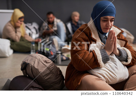Portrait of adult Middle Eastern woman wearing hijab and praying in refugee shelter, copy space 110640342