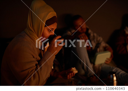 Diverse group of Middle Eastern people hiding in dark bomb shelter with young woman drinking hot tea in foreground Diverse group of Middle Eastern people hiding in dark bomb shelter with young woman drinking hot tea in foreground 110640343