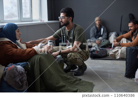 Group of Middle Eastern people sitting on floor in refugee shelter focus on doctor talking to young woman in foreground, copy space 110640386