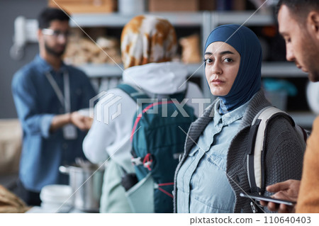 Waist up portrait of adult Middle Eastern woman wearing hijab and looking at camera while standing in line at refugee help center, copy space 110640403