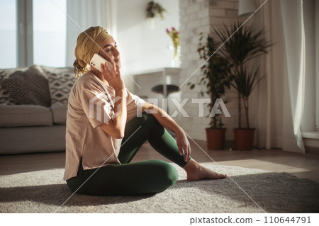Young woman with cancer sitting on floor, phone calling in her apartment. Strong female patient talking with friend, family on phone, feeling loved. Young woman with cancer sitting on floor, phone calling in her apartment. Strong female patient talking with friend, family on phone, feeling loved. 110644791