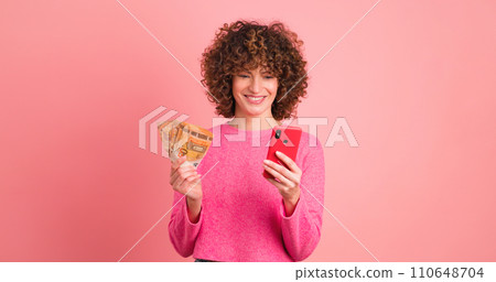 Excited woman with cash and looking mobile phone in pink studio 110648704