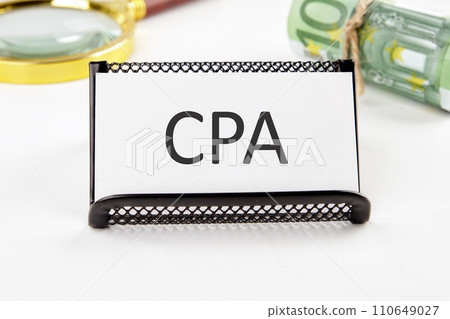 CPA, Certified Public Accountant or Cost per Action the text on the business card next to the roll of money with a magnifying glass in the background is out of focus on a white background 110649027
