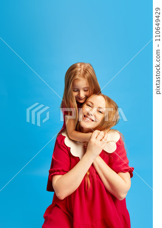 Portrait of beautiful young mother posing with her little daughter in same color dresses against blue studio background. Mother's Day, International Happiness Day 110649429