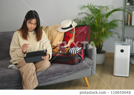 Young asian woman booking hotel online on digital tablet preparing stuff and clothes for holiday, vacation trip at home. Young asian woman booking hotel online on digital tablet preparing stuff and clothes for holiday, vacation trip at home. 110650048
