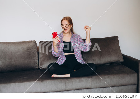 Satisfied young woman holding phone and celebrating on sofa at home. Smiling female sitting cross-legged on comfortable couch, happy lady relaxing with mobile phone in cozy living room, full body Satisfied young woman holding phone and celebrating on sofa at home. Smiling female sitting cross-legged on comfortable couch, happy lady relaxing with mobile phone in cozy living room, full body 110650197