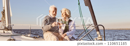Good vibes. Happy senior couple sitting on the side of sail boat or yacht deck floating in sea. Man and woman drinking wine or champagne, enjoying the view 110651185