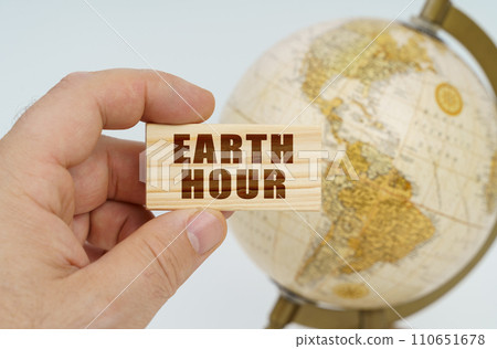 A man holds in his hand a wooden plate on which it is written - Earth Hour. In the background is a globe. A man holds in his hand a wooden plate on which it is written - Earth Hour. In the background is a globe. 110651678