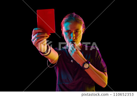 Young concentrated woman, soccer referee blowing a whistle and showing red card as dismissal symbol against black studio background in neon light Young concentrated woman, soccer referee blowing a whistle and showing red card as dismissal symbol against black studio background in neon light 110652353