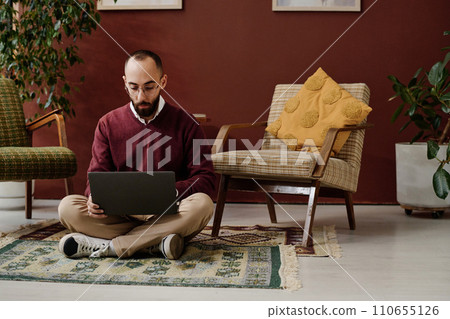 Middle eastern man in casual outfit sitting cross-legged on floor in office and browsing his laptop Middle eastern man in casual outfit sitting cross-legged on floor in office and browsing his laptop 110655126