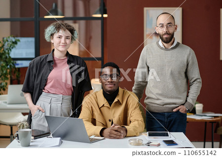 Two biracial male coworkers and their female colleague posing by office desk smiling at camera 110655142