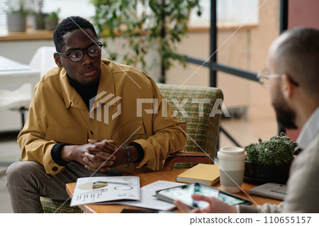 Black financial specialist sitting in armchair at office listening to his colleague 110655157