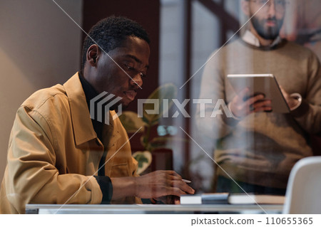 Black man in casual outfit working at desk in office, his caucasian colleague standing on background 110655365