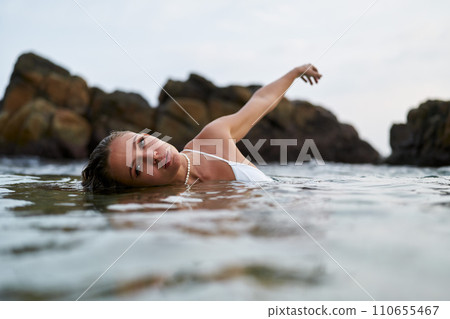 Woman swims gracefully in tranquil lagoon wearing waterproof cosmetics during golden hour. Fashionable swimwear, serene nature backdrop ensures leisure. Female enjoys secluded dip in serene waters. 110655467
