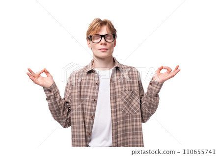 a young caucasian red-haired guy in a shirt over a t-shirt meditates with his eyes closed 110655741