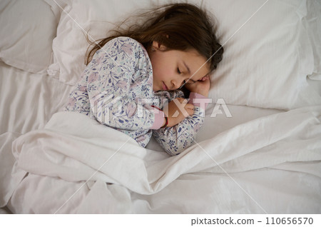 Top view beautiful lovely little child girl in stylish pajamas, sleeping on her bed, covered with white clean bedsheets Top view beautiful lovely little child girl in stylish pajamas, sleeping on her bed, covered with white clean bedsheets 110656570