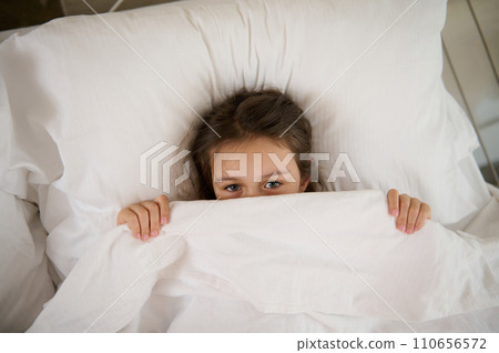 Top view of an adorable child girl lying on the bed, hiding half of her face under white blanket, looking at the camera Top view of an adorable child girl lying on the bed, hiding half of her face under white blanket, looking at the camera 110656572