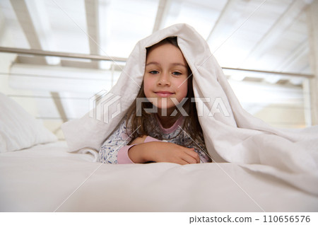 Close-up portrait of a little child girl 6 years old lying on the bed under white blanket while waking up in the morning Close-up portrait of a little child girl 6 years old lying on the bed under white blanket while waking up in the morning 110656576