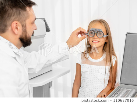 Doctor ophthalmologist holding special eye equipment examinating girl's eyes in the clinic Doctor ophthalmologist holding special eye equipment examinating girl's eyes in the clinic 110657016