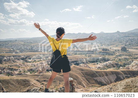 Woman tourist with backpack standing backwards on mountain top in Cappadocia with open arms, Turkey 110657023