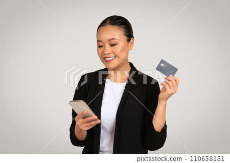 Smiling businesswoman in a black suit holding a smartphone in one hand and a credit card 110658181