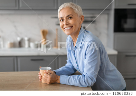 Smiling senior lady stands by table savoring morning coffee indoor Smiling senior lady stands by table savoring morning coffee indoor 110658383
