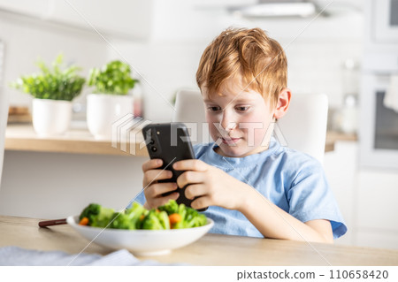 View of smart cute boy using smartphone before lunch in kitchen at home View of smart cute boy using smartphone before lunch in kitchen at home 110658420