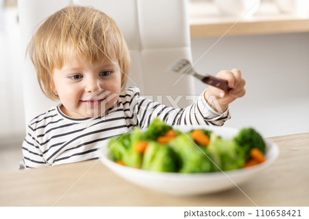 Cute little girl is eating broccoli and carrot vegetables with a fork Cute little girl is eating broccoli and carrot vegetables with a fork 110658421