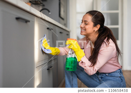 Smiling young woman cleaning kitchen cabinet with spray and cloth Smiling young woman cleaning kitchen cabinet with spray and cloth 110658438