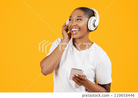black lady using phone listening music wearing earphones, yellow background 110658446
