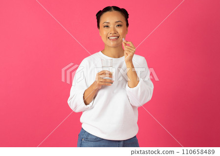 Happy Young Asian Lady Holding Glass Of Water And Beauty Supplement Pill 110658489