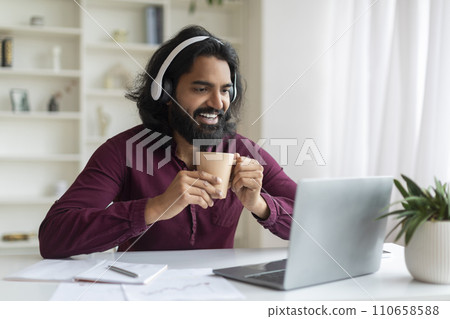 Indian man enjoying coffee break while working on laptop in home office Indian man enjoying coffee break while working on laptop in home office 110658588