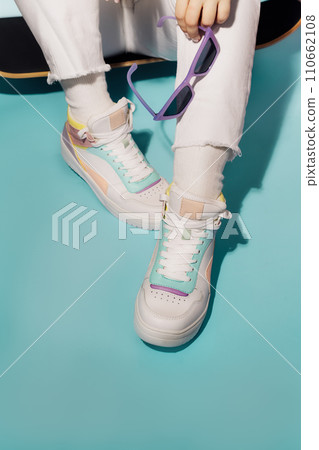 Close up female legs in white jeans and retro style high-top multicolor sport sneakers shoes sitting on the skateboard on turquoise blue background. Vintage retro fashion style of 80s - 90s vibes Close up female legs in white jeans and retro style high-top multicolor sport sneakers shoes sitting on the skateboard on turquoise blue background. Vintage retro fashion style of 80s - 90s vibes 110662108