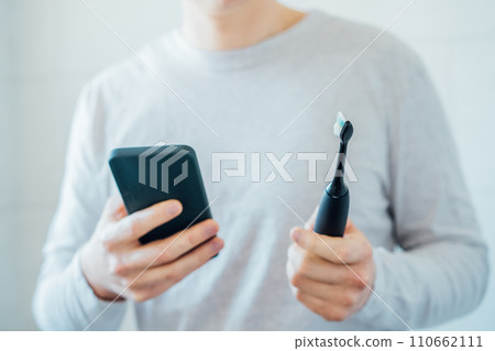 Close up man puts settings of his electronic toothbrush with mobile phone app. Wireless connecting sonic toothbrush with smart phone app. Modern home health care technology concept. Selective focus 110662111