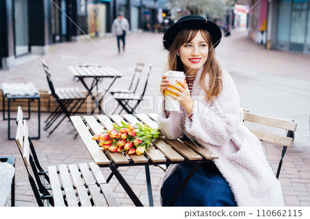 Stylish young smiling woman enjoying coffee from a reusable cup, listening to music, podcast using wireless earbuds and phone in street cafe. Fresh tulips bouquet on table. Springtime street fashion. Stylish young smiling woman enjoying coffee from a reusable cup, listening to music, podcast using wireless earbuds and phone in street cafe. Fresh tulips bouquet on table. Springtime street fashion. 110662115