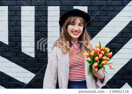 Portrait of happy. smiling young fashion woman holding bouquet of fresh tulip flowers on the painted brick wall background. Urban city street fashion. Spring positive mood. Selective focus Portrait of happy. smiling young fashion woman holding bouquet of fresh tulip flowers on the painted brick wall background. Urban city street fashion. Spring positive mood. Selective focus 110662116